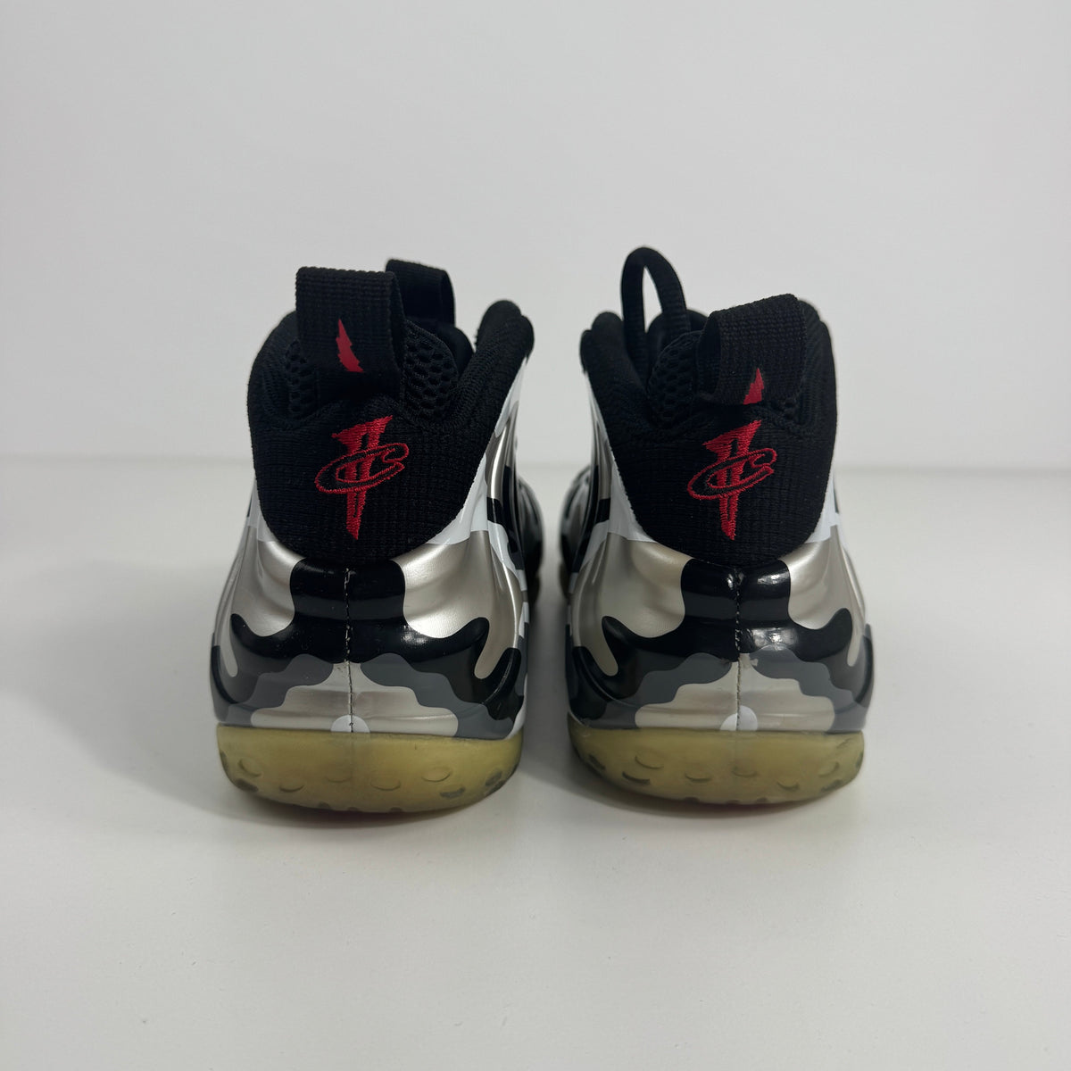 NIKE Air Foamposite One trainers viewed from the back, with a shiny silver and black design, red logo pull tabs and yellowed soles.