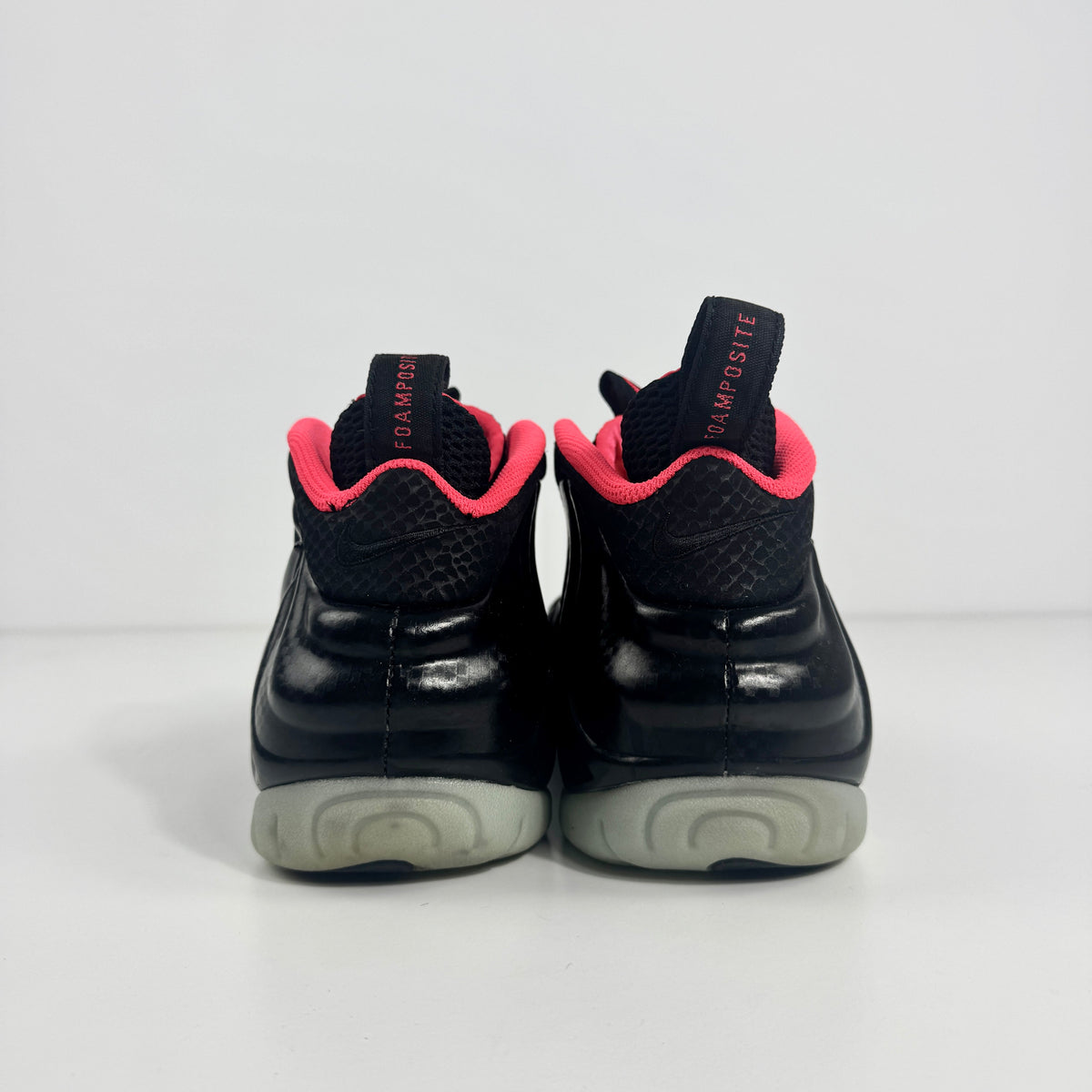 Back view of black Nike trainers with a pink inner lining, grey soles, and black pull tabs featuring red text, on a white background.